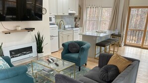 A modern living room with a sofa, armchair, and coffee table. A kitchen with white cabinets and appliances is visible in the background. - Glasshouse in the Woods. Five acres of wooded privacy and wildlife. (Augusta)