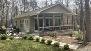 A small, single-story cabin with a covered porch and a patio area. - Glasshouse in the Woods. Five acres of wooded privacy and wildlife. (Augusta)