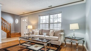 A living room with a sofa, coffee table, and two lamps. There is a window with a view of trees and a door with a window. - Indianapolis Home w/ Private Yard ~ 8 Mi to Dtwn! (Indianapolis)