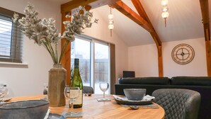 A dining area with a round wooden table, a vase with flowers, a wine bottle, and a black leather couch. - The Stables @ Merrydown (Axminster)