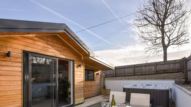 A wooden cabin with a glass door entrance, a patio with wicker furniture, and a hot tub. - The Stables @ Merrydown (Axminster)