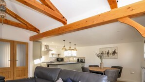 A modern living room with a grey leather sofa, wooden beams, a dining area with a table and chairs, and a kitchen in the background. - The Stables @ Merrydown (Axminster)