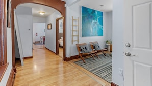 A hallway with wooden flooring, a large abstract painting on the wall, a ladder, and a bench with pillows.