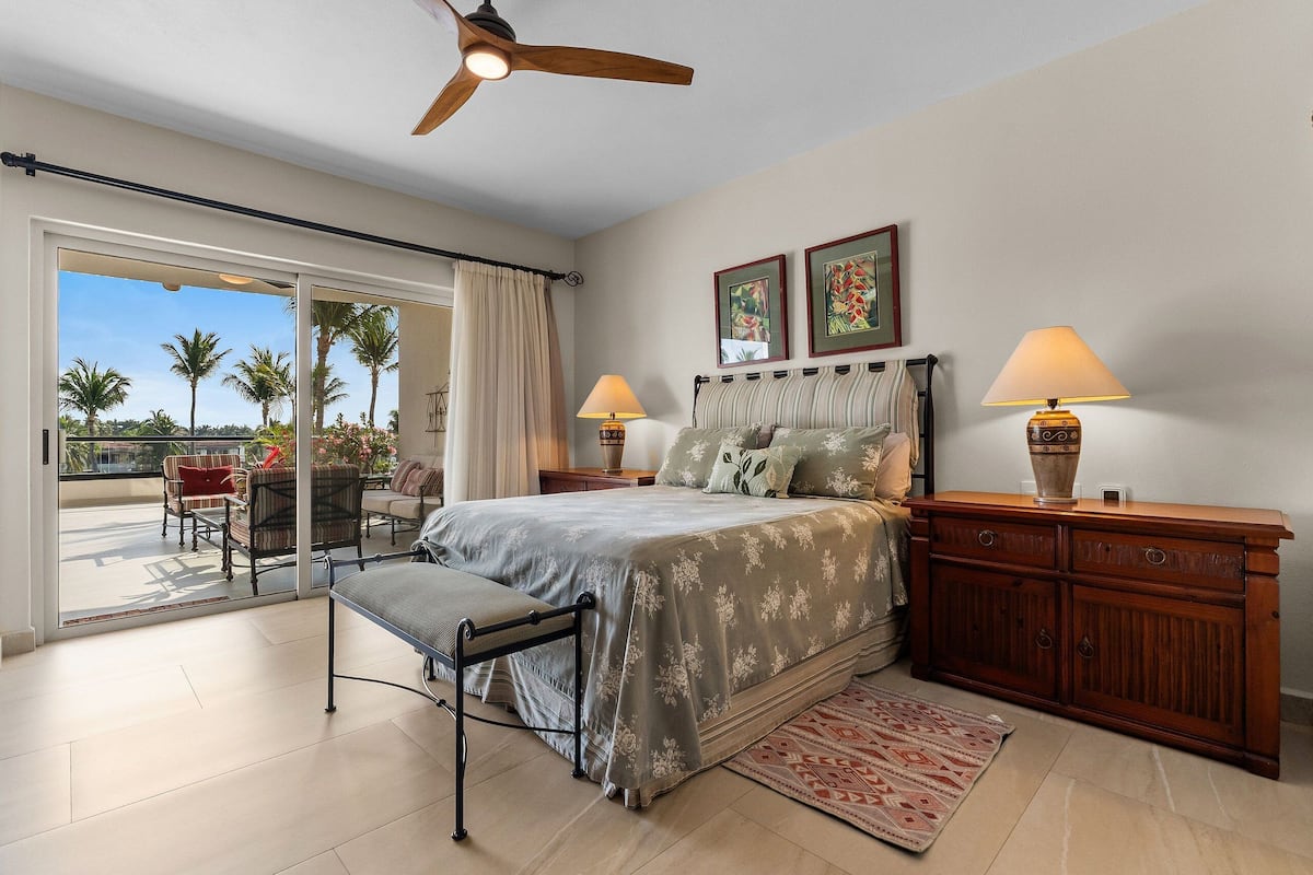 A bedroom with a bed, a bench, a nightstand, a lamp, a ceiling fan, and a view of palm trees through a sliding door.