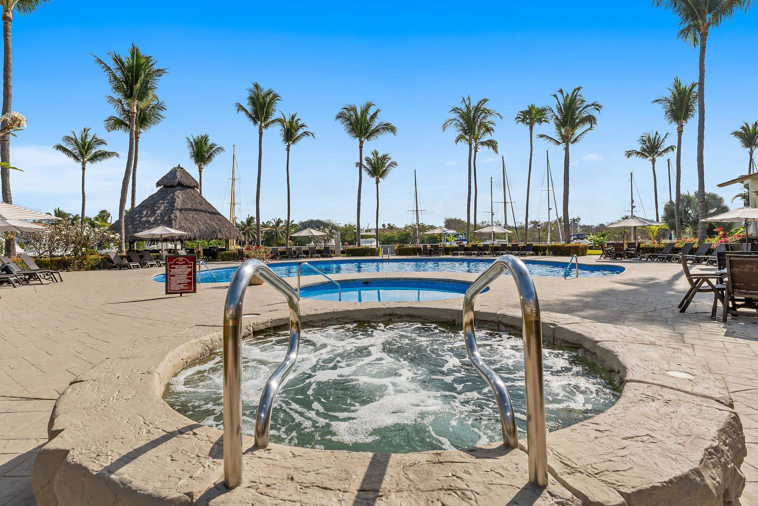 A swimming pool with a hot tub, surrounded by palm trees and lounge chairs.