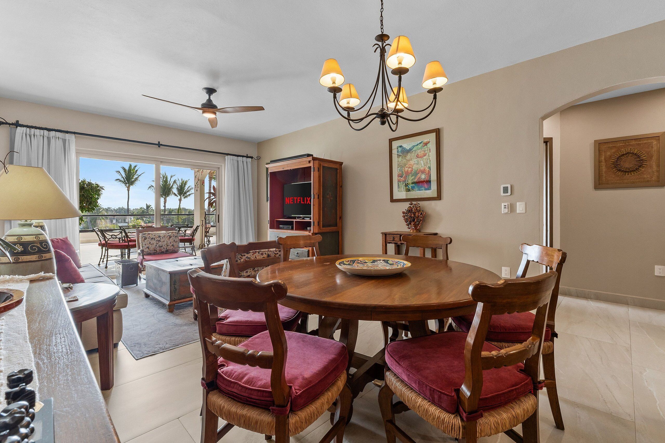 A dining area with a round table, wooden chairs with red cushions, a chandelier, and a view through large windows.
