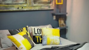 Un lit bien fait, recouvert d’une couverture grise, avec deux oreillers aux motifs jaunes et noirs, et une serviette blanche enroulée.