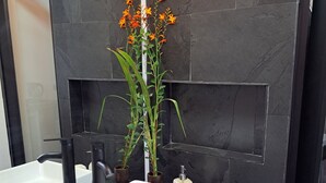 Une salle de bain moderne avec un mur en marbre noir, deux vasques blanches, un miroir et une plante en pot aux fleurs orange.