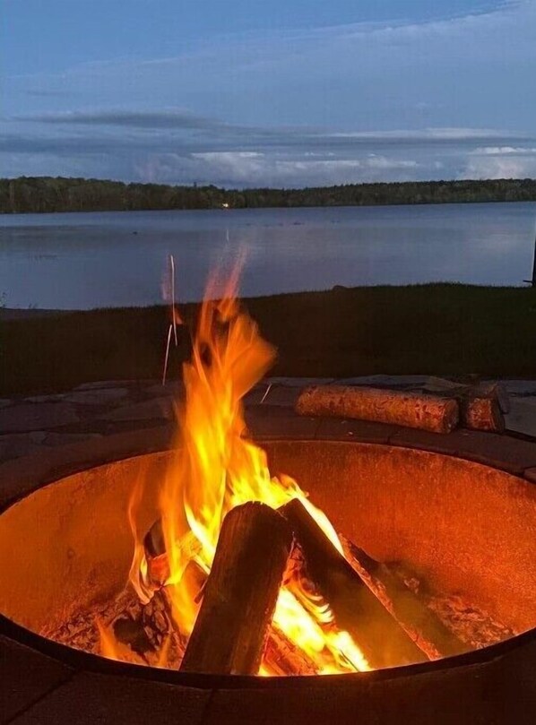 Un foyer avec des bûches, entouré d’une bordure en pierre, donnant sur un lac paisible au crépuscule.
