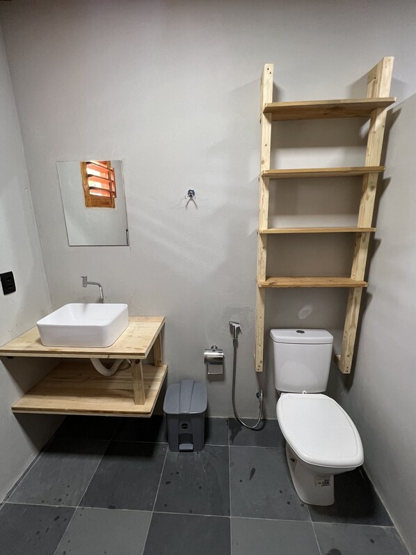 A compact bathroom with a white sink, a toilet, and a wooden shelf unit. - Windland (Amontada)