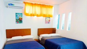 Une chambre avec deux lits en bois, chacun recouvert d’une courtepointe matelassée bleue et agrémenté d’oreillers blancs. Il y a une fenêtre donnant sur le ciel, ainsi qu’un climatiseur fixé au mur.