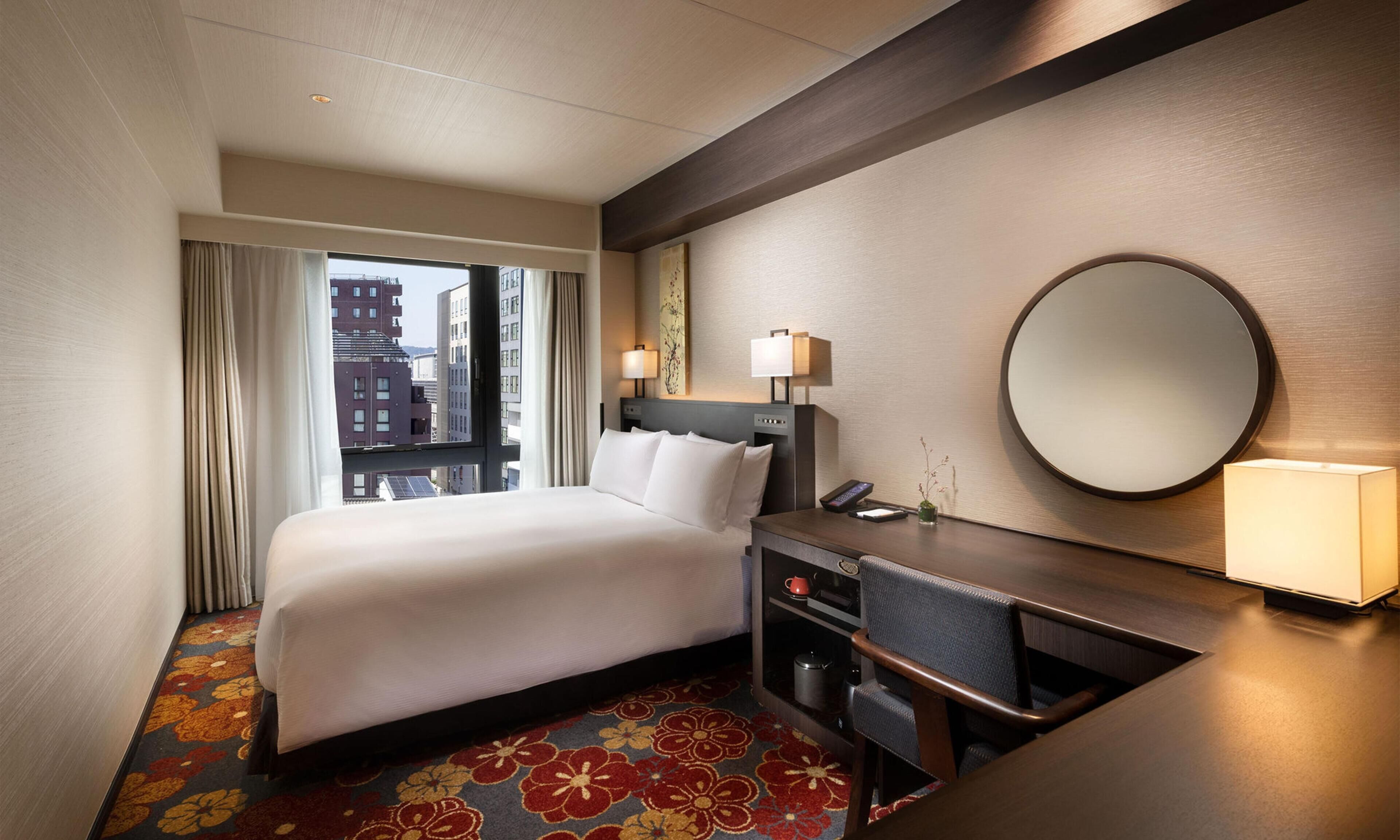 Een hotelkamer met een bed, bureau, spiegel en uitzicht op gebouwen.