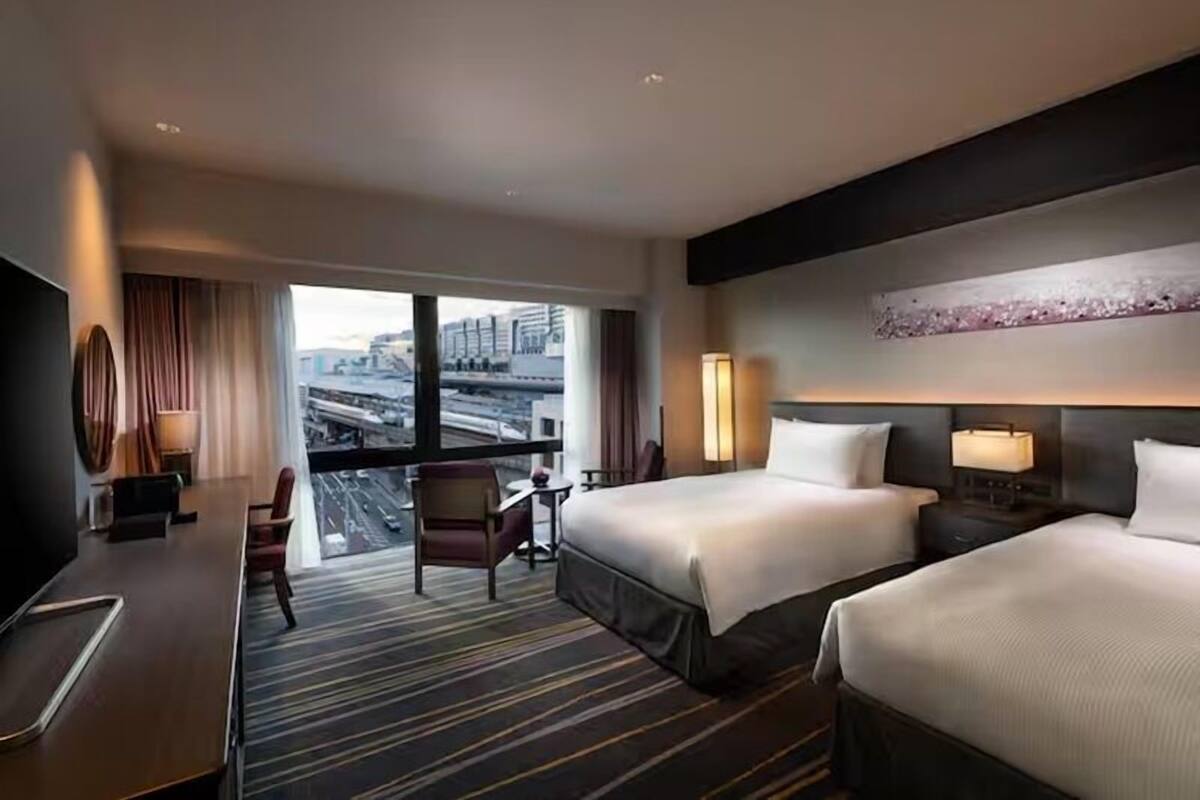 A hotel room with two beds, a desk, a chair, a TV, and a large window offering a city view.