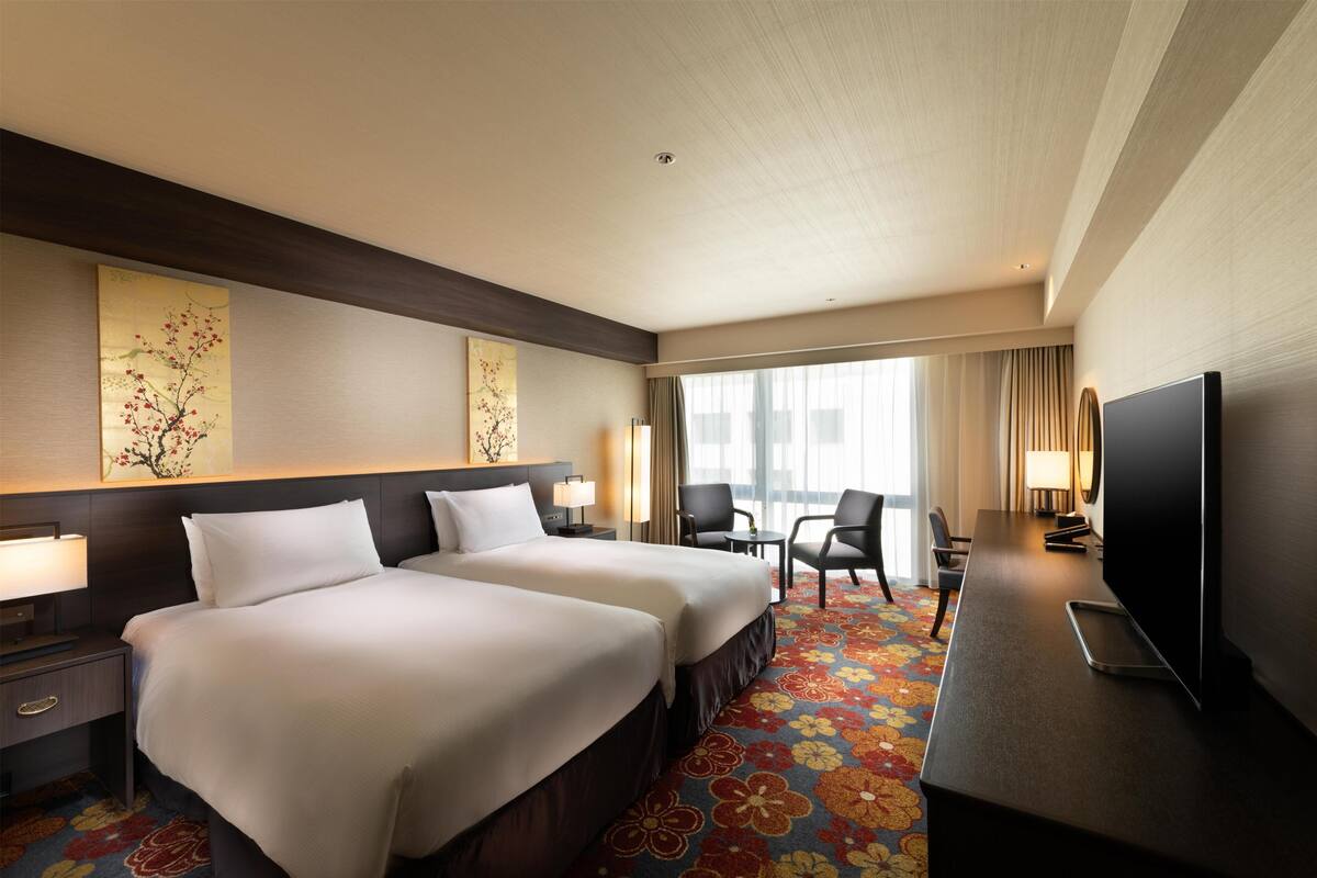 A hotel room with two beds, a TV, a desk, and a large window.