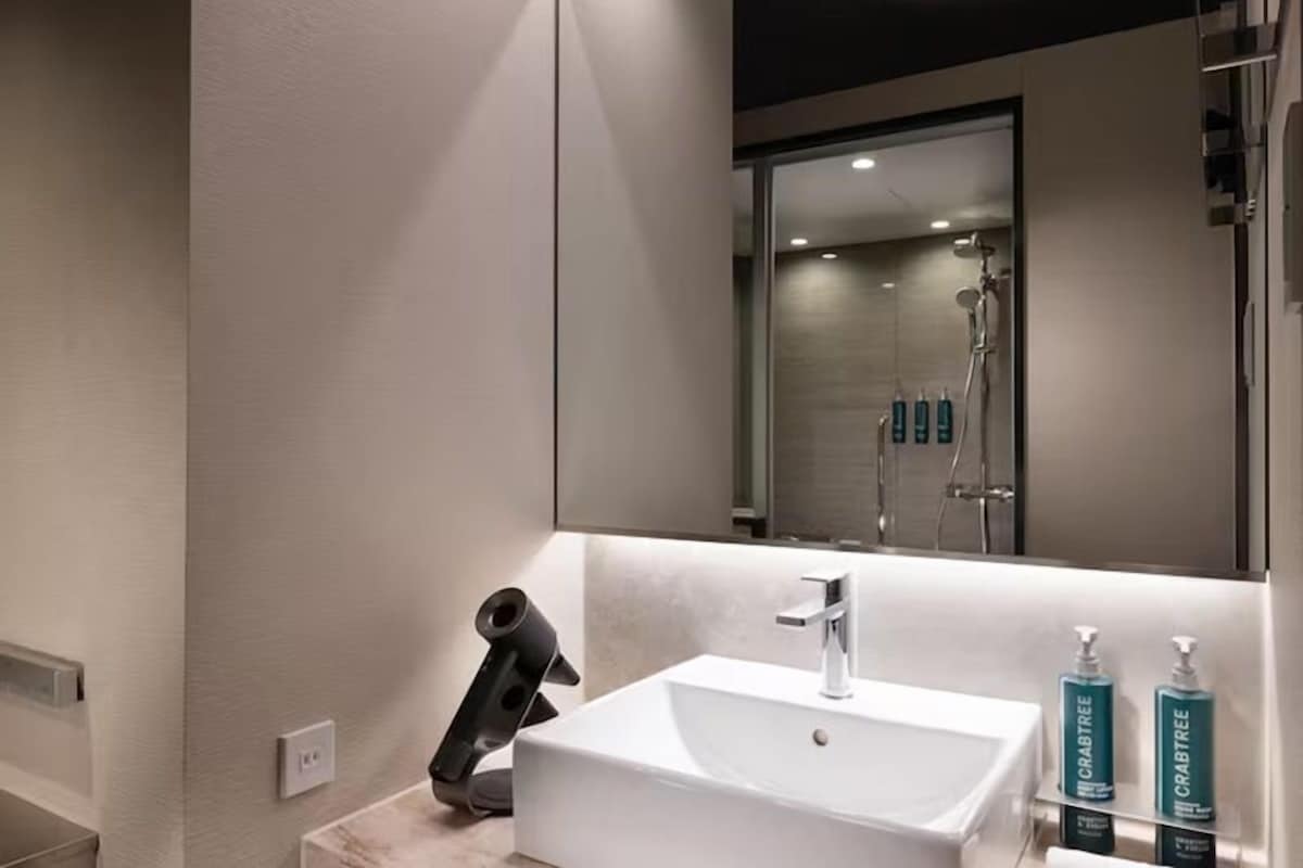A modern bathroom with a white sink, a mirror, and a shower visible in the reflection.