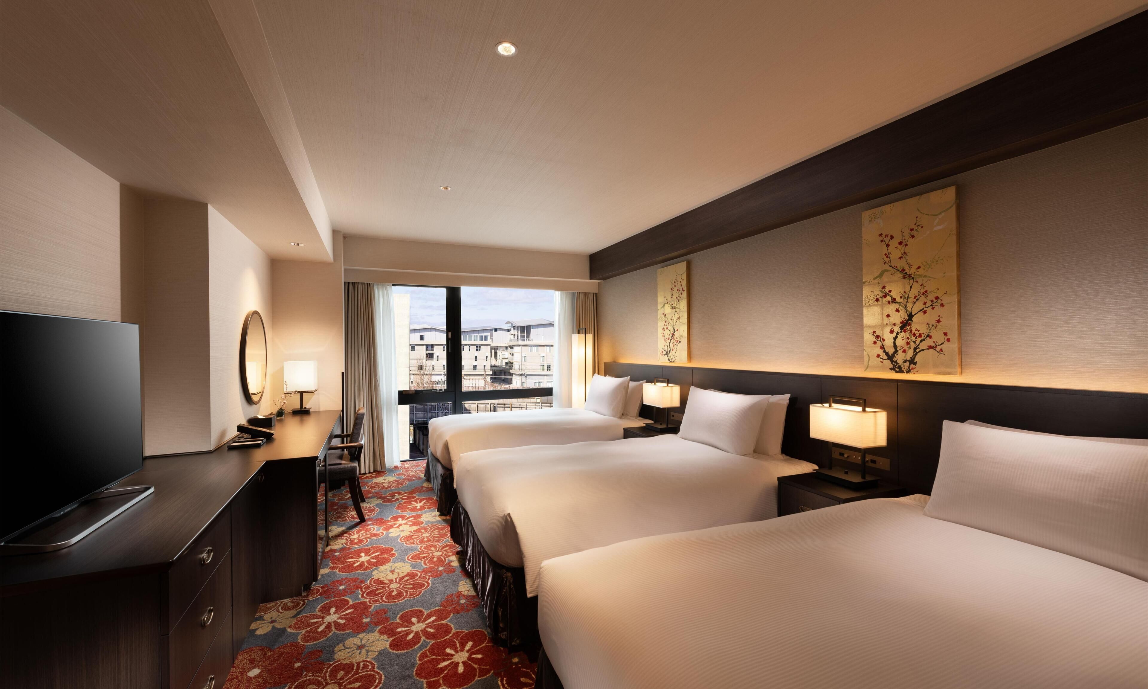Een hotelkamer met twee bedden, een bureau met een televisie, een groot raam en uitzicht op gebouwen.