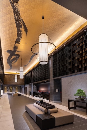 Een ruime lobby met een hoge plafond, moderne hanglampen, een receptiebalie en een zithoek met een potplant.