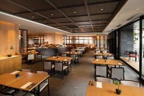 Um restaurante moderno com mesas e cadeiras de madeira, uma área de bar central e grandes janelas com vista para o exterior.