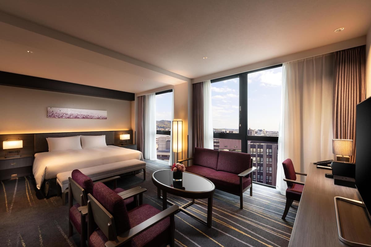 A hotel room with a bed, a sofa, a desk, a TV, and large windows.