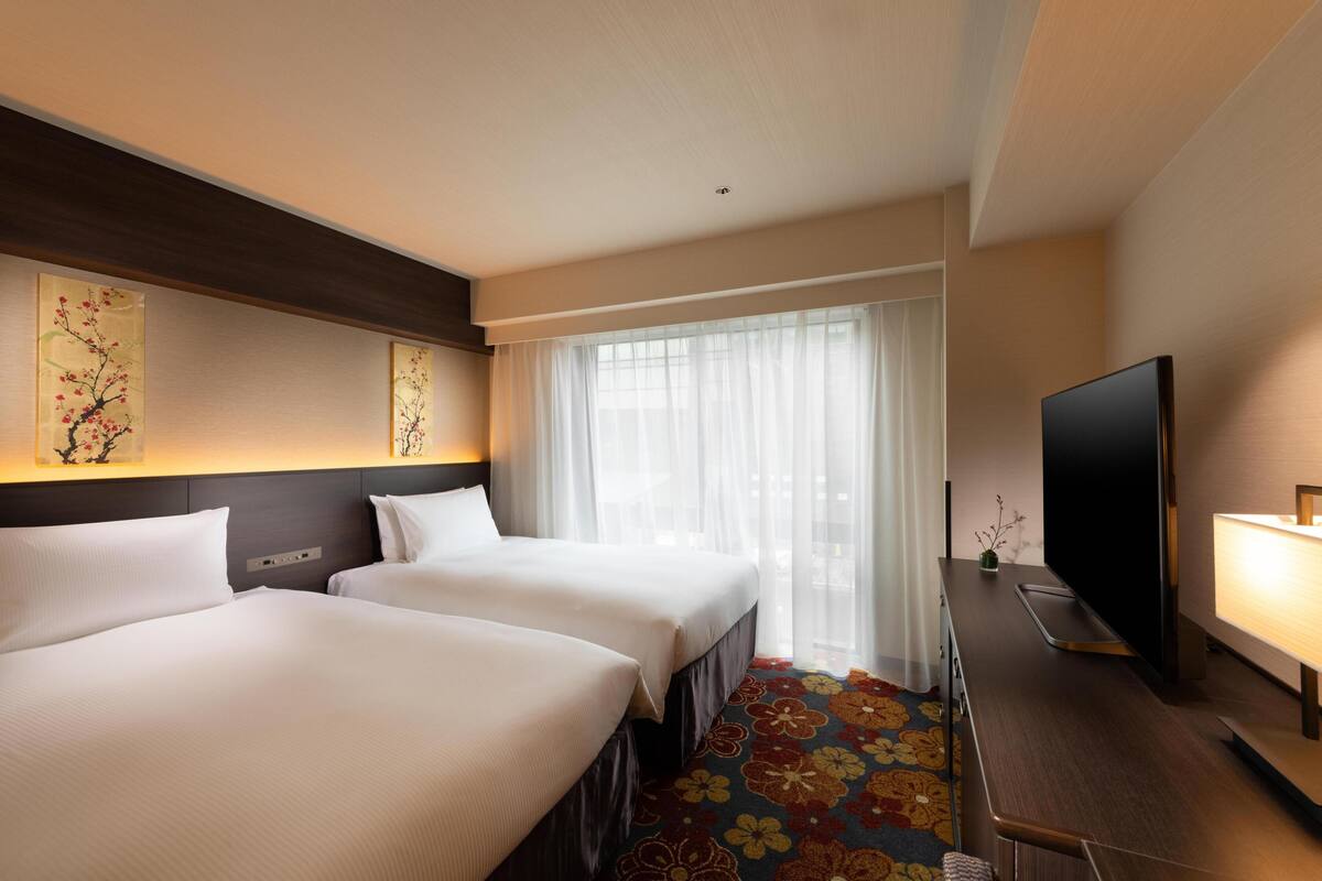 A hotel room with two beds, a TV, a desk with a lamp, and a large window with sheer curtains.