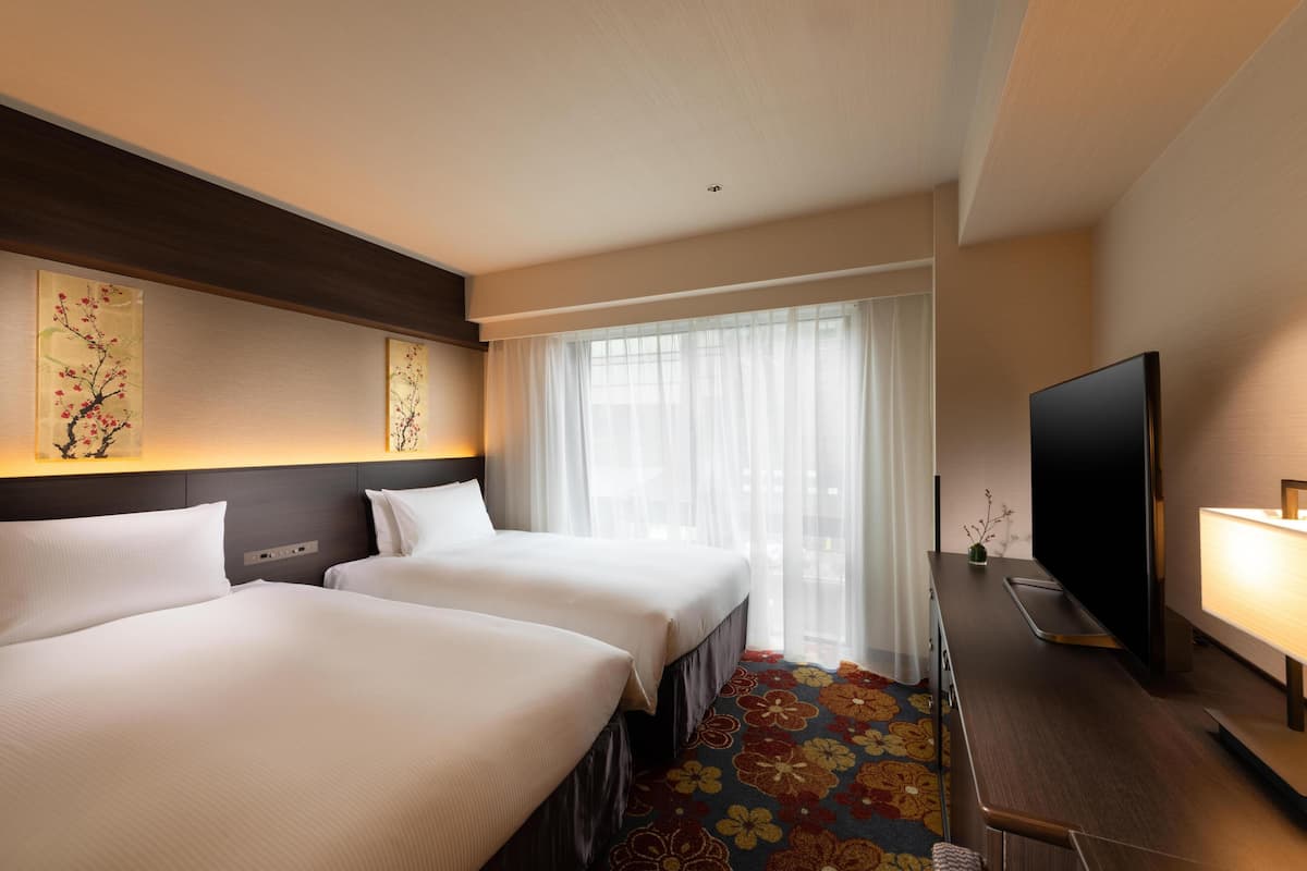 Een hotelkamer met twee bedden, een televisie, een bureau met lamp en een groot raam met dunne gordijnen.