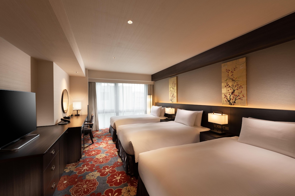 A hotel room with a TV, three beds, a desk, and a large window.
