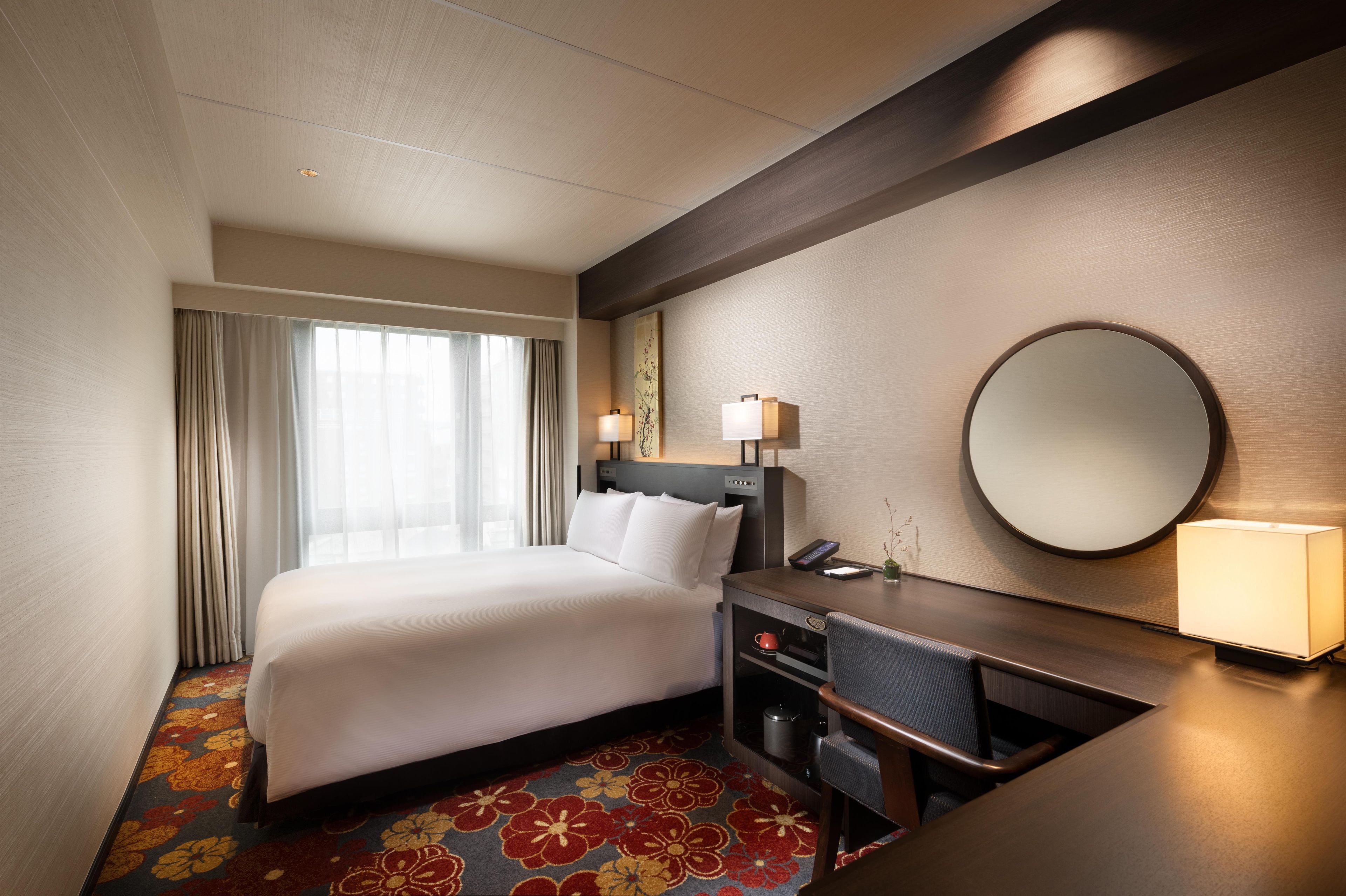 A hotel room with a bed, desk, chair, desk lamp, and a round mirror.