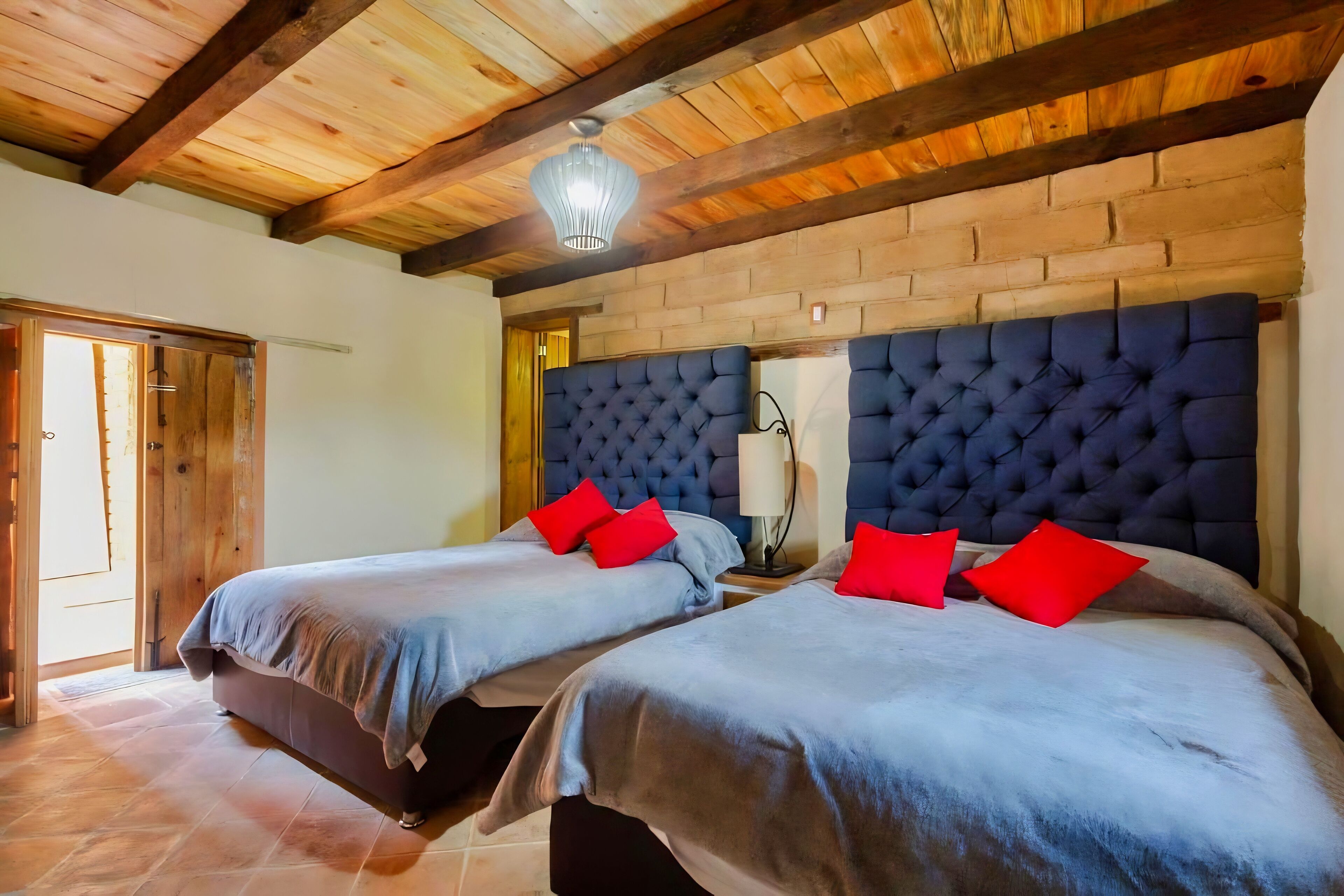 A room with two beds, each with a headboard and red pillows, under a wooden ceiling with exposed beams.