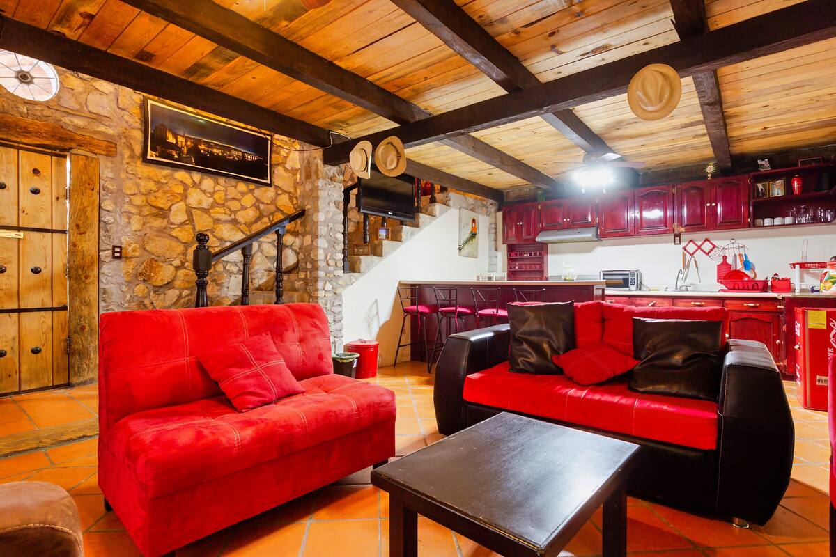 A cozy living area with a red sofa, a black armchair, and a wooden coffee table. A stone wall and wooden beams are visible, along with a kitchen area in the background.