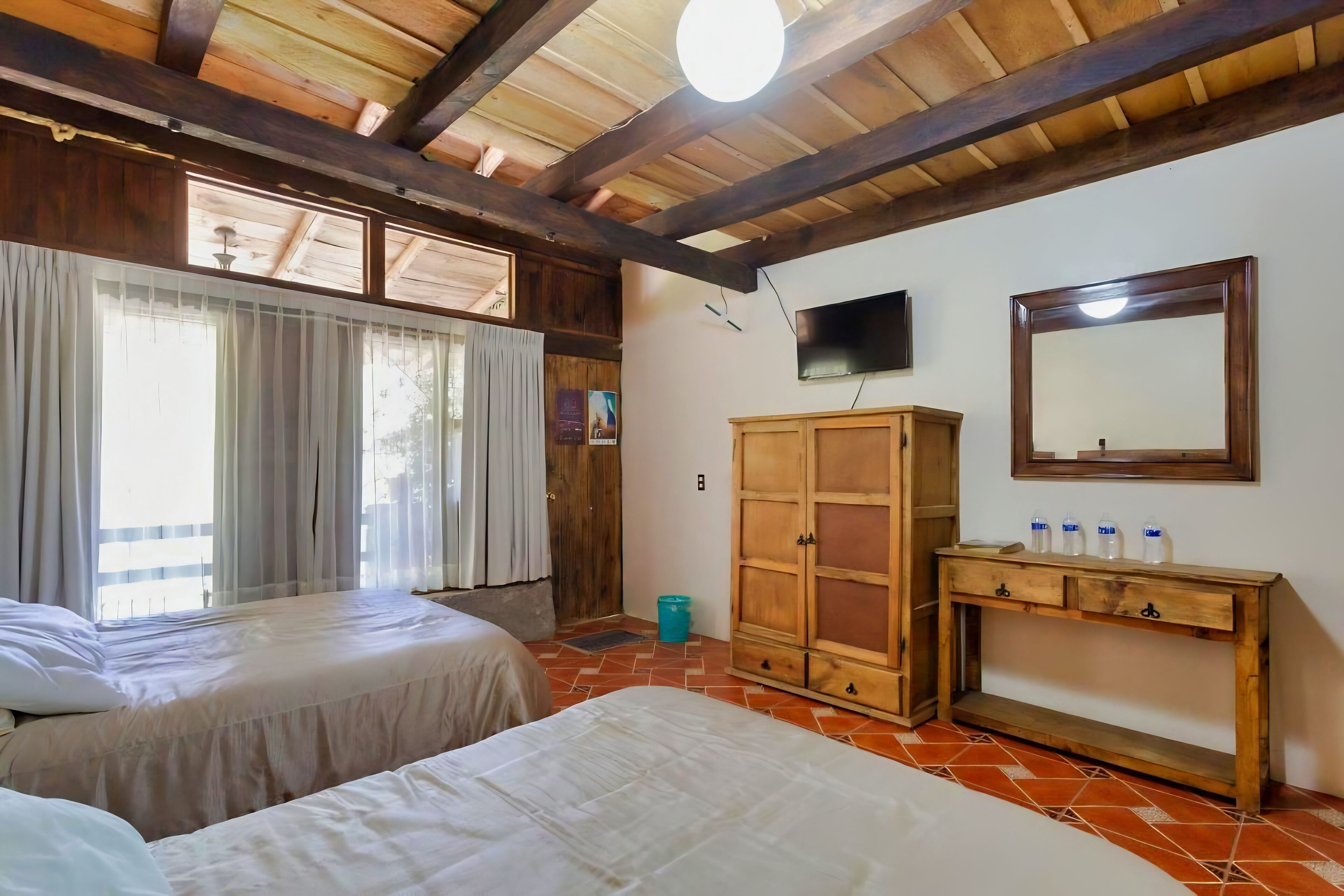 A room with two beds, wooden ceiling beams, a wooden cabinet, a television, and a tiled floor.