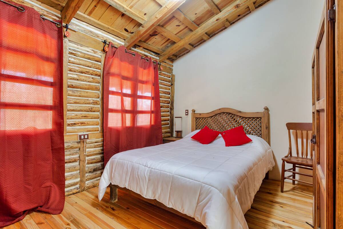 A wooden cabin-style bedroom with a bed, red curtains, and a wooden chair.