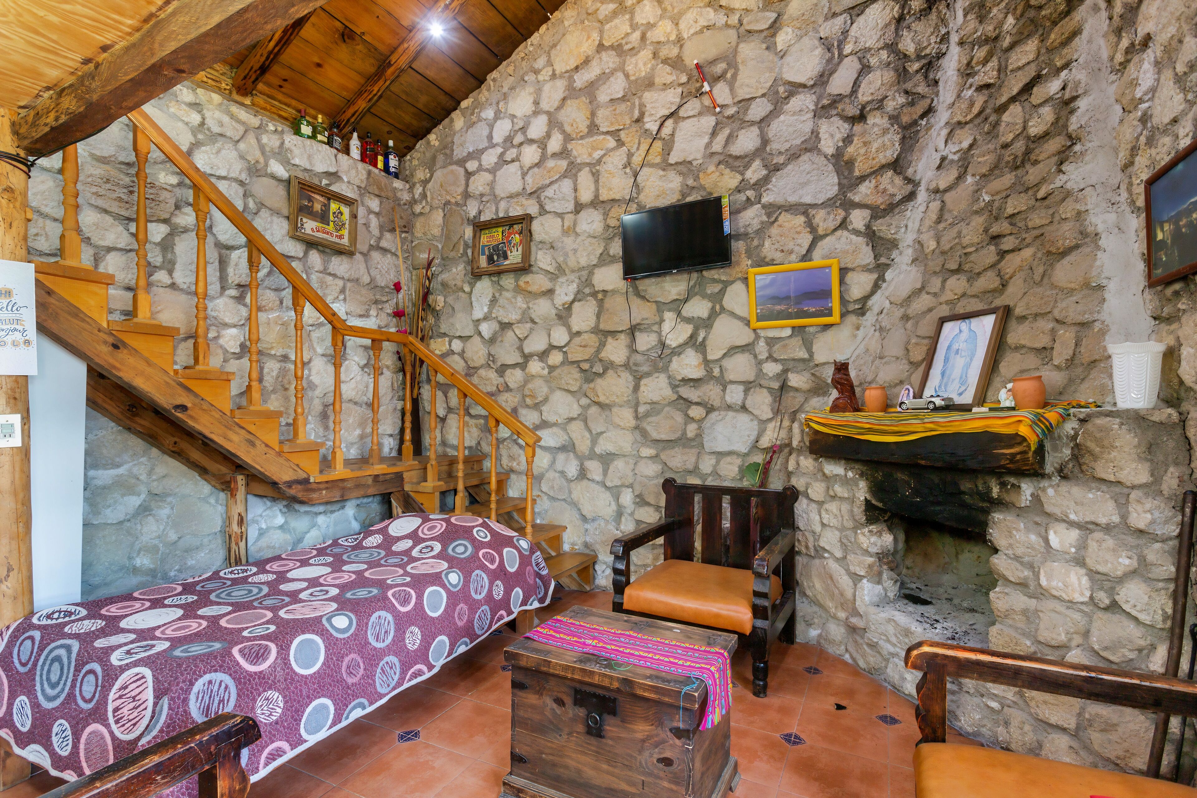 Una habitación rústica con pared de piedra, escalera de madera, cama con colcha estampada, un cofre de madera, una silla y televisor montado en la pared.