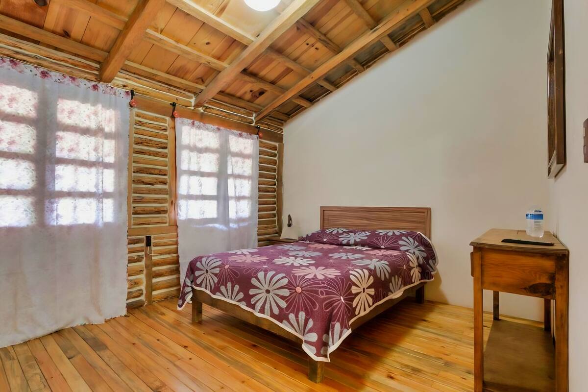 A wooden-paneled room with a bed, a small table, and a window with curtains.
