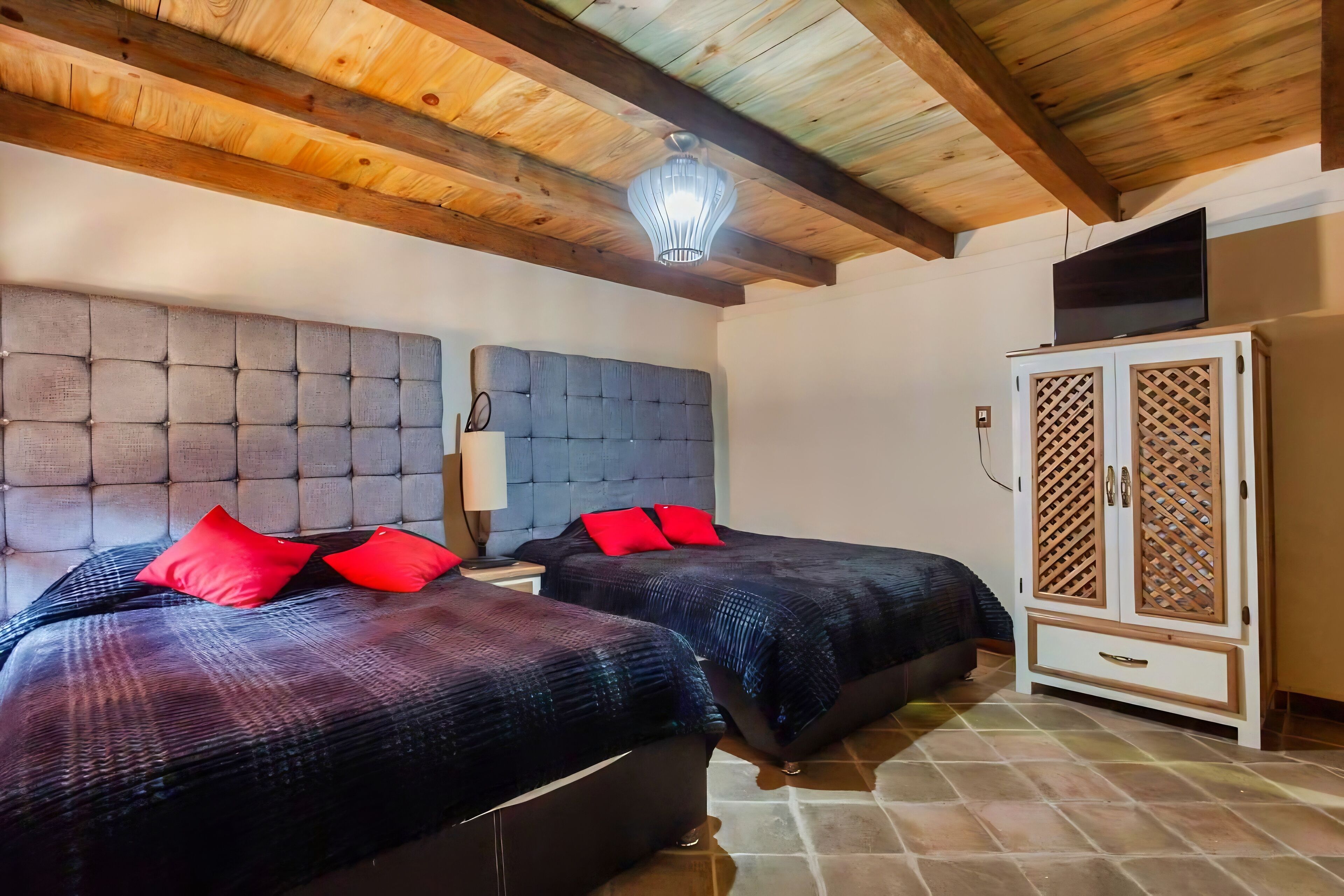 A bedroom with two beds, a wooden ceiling, a TV on a cabinet, and a nightstand with a lamp.