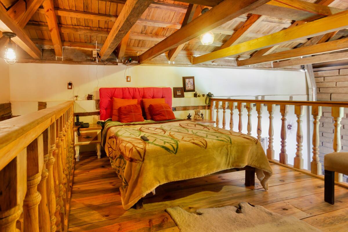 A wooden-floored bedroom with a red headboard, a bed with a green bedspread, and a wooden railing along the balcony.