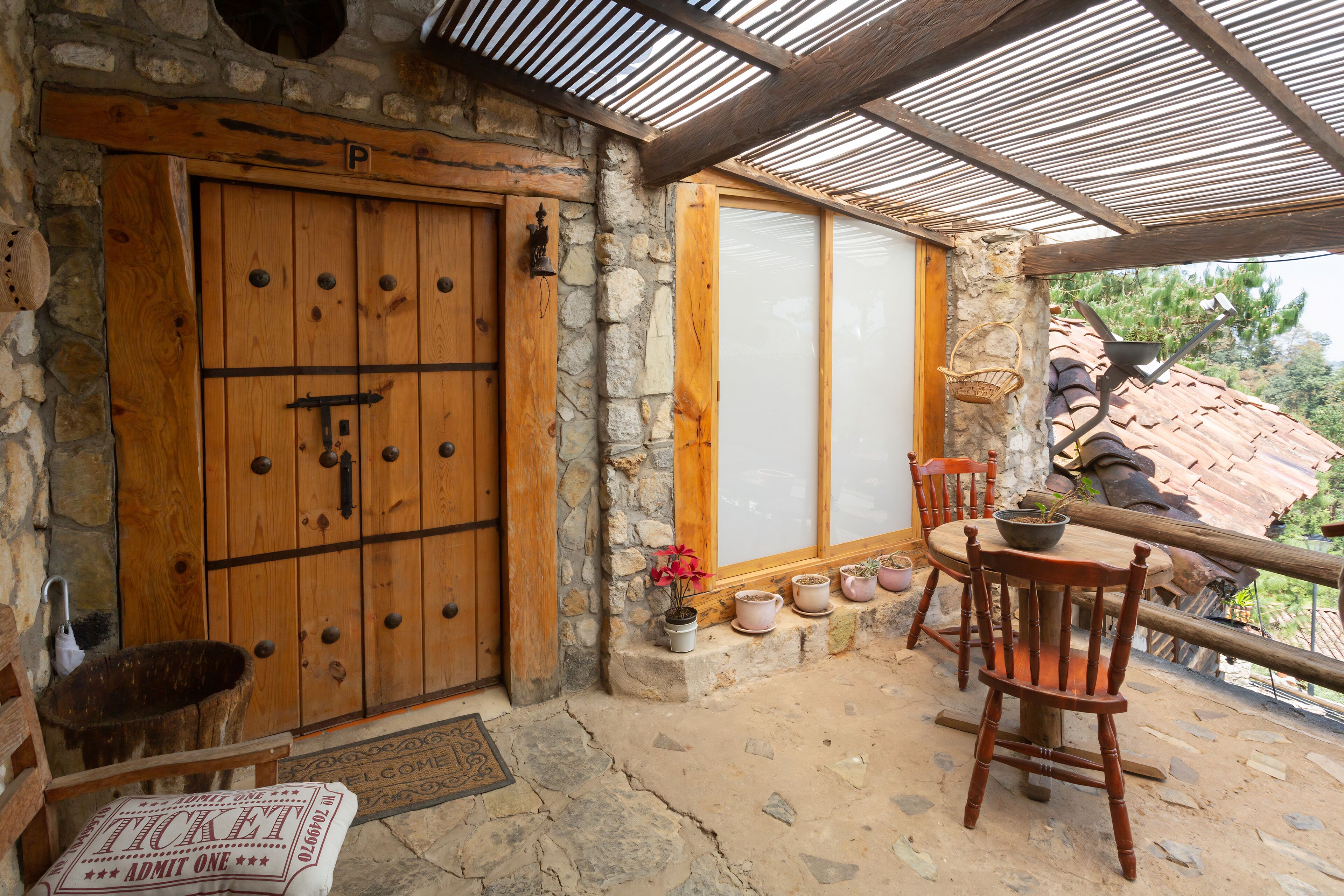A rustic stone building with a wooden door, a covered patio with a table and chairs, and potted plants.