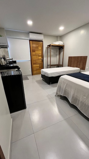A compact hotel room with two single beds, a wooden wardrobe, a small desk with a chair, and a kitchenette area.