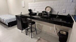 Cozinha compacta com bancada preta, painel de azulejos brancos, pia pequena e uma cadeira.