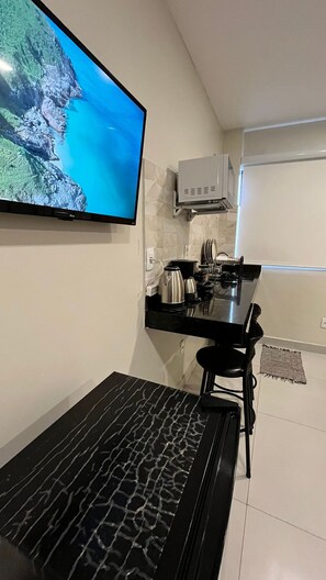A compact kitchen with a flat-screen TV mounted on the wall, a small countertop with a kettle, and a black chair.