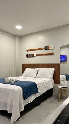 Quarto de hotel com cama, almofadas, manta azul, cabeceira de madeira, televisão de tela plana e uma mesinha com controle remoto.