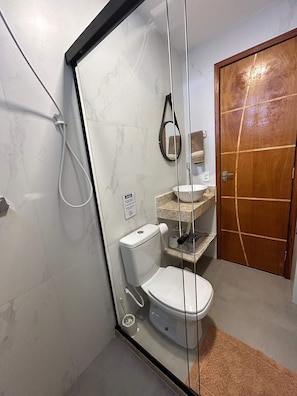 A modern bathroom with a glass shower enclosure, a white toilet, a sink with a marble countertop, and a wooden door. - Morada do Sol Suítes - Suíte 04 (Arraial do Cabo)