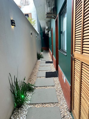A narrow pathway with stepping stones, bordered by a white wall and a wooden door on one side. - Morada do Sol Suítes - Suíte 04 (Arraial do Cabo)