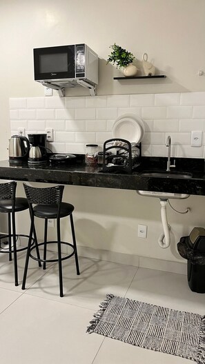 Una cocina con encimera negra, salpicadero blanco de azulejos, microondas, fregadero, dos sillas y una pequeña planta en maceta.