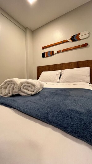 Una cama bien hecha con una manta azul y una toalla blanca enrollada, cabecero de madera y dos esquíes de madera apoyados contra la pared.