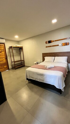 A hotel room with a bed, a wooden wardrobe, a small table, and a wall-mounted shelf. - Morada do Sol Suítes - Suíte 05 (Arraial do Cabo)