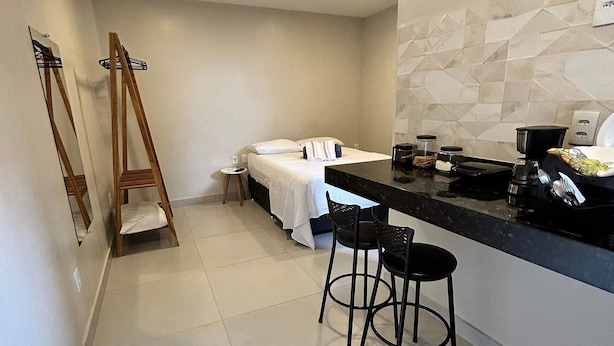 A modern hotel room with a bed, a small sitting area with two chairs, a kitchen counter, and a large mirror. - Morada do Sol Suítes - Suíte 06 (Arraial do Cabo)
