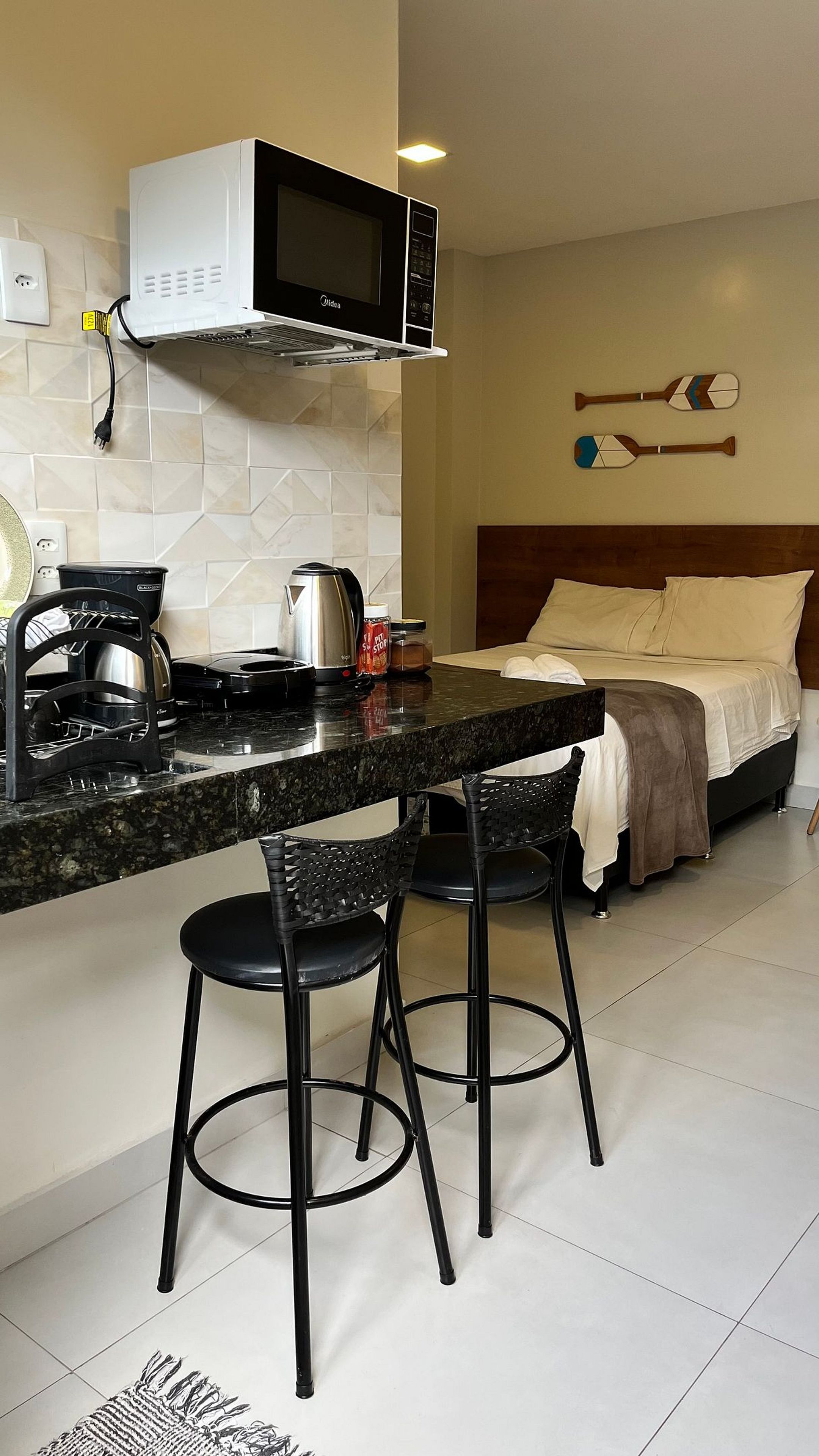 Cozinha compacta com micro-ondas, chaleira e máquina de café. Há dois bancos altos e uma cama com cabeceira e almofadas.