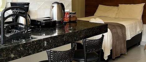 Cocina compacta equipada con microondas, hervidor y cafetera. Hay dos taburetes de bar y una cama con cabecera y almohadas.