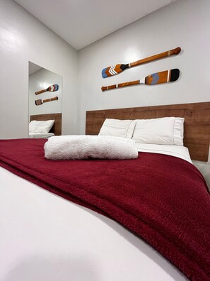 Cama bem arrumada com manta vermelha e almofadas brancas, cabeceira de madeira e dois remos decorativos pendurados na parede.