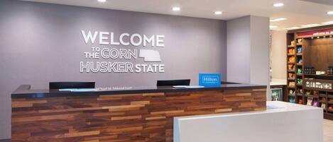 A hotel reception desk with a "WELCOME TO THE CORN HUSKER STATE" sign, a Hilton Honors badge, and a display of amenities.